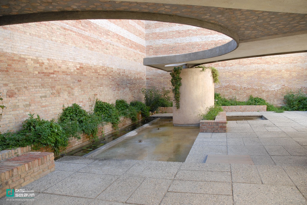 نمایی آرام از «Giardino delle Sculture» کارلو اسکارپا در بینال ونیز؛ حیاطی محصور با دیوارهای آجری، حوضچه آب خطی، گیاهان سبز در پایه دیوارها و ستون استوانه‌ای پوشیده از گیاه، زیر سقف بتنی منحنی با بازشوی دایره‌ای.